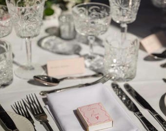 Puede incluir: Una pequeña caja de cerillas rectangular de color rosa con bordes dorados sobre una servilleta blanca doblada. La caja presenta un borde ondulado rosa, el texto manuscrito "Claire & Noel" y un pequeño corazón. Cubiertos de plata y vasos la rodean.