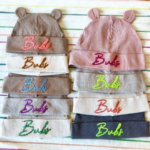 May include: A stack of eight knitted hats in various colors with the word "Bub" embroidered on each hat in different colors.