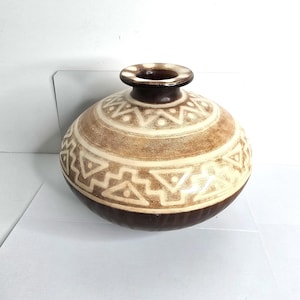 May include: A decorative ceramic vase with a rounded body and a narrow neck. The vase features a brown base, a cream-colored middle section with geometric patterns, and a dark brown rim. The overall design is earthy and textured.