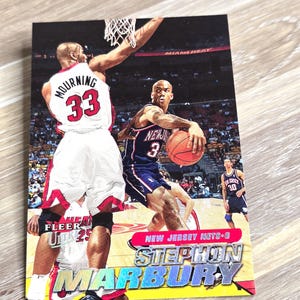 May include: A basketball trading card featuring Stephon Marbury of the New Jersey Nets. The card shows Marbury dribbling a basketball, with Alonzo Mourning in the background. The card has the Fleer logo and the text "NEW JERSEY NETS-6".