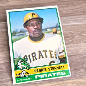 May include: A vintage baseball card featuring Rennie Stennett of the Pittsburgh Pirates. The card has a white background with green and yellow accents. Stennett is wearing a yellow helmet and a white jersey with the word "Pirates".