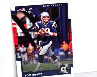 May include: A Tom Brady football card featuring the New England Patriots. The card has a white border with a blue and red design. Brady is wearing a blue and white uniform and holding a football. The Donruss logo is visible.