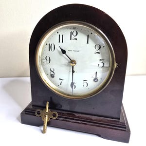 May include: A dark brown wooden mantel clock with an arched top and a white circular face featuring black Arabic numerals. The dial displays the text Seth Thomas. A brass winding key rests at the base of the clock, which has black metal hands.