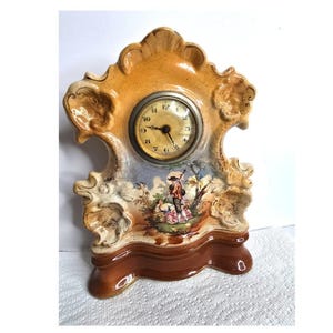 May include: A decorative ceramic clock with a cream-colored, ornate frame and a brown base. The clock face is round with black numerals. The scene on the clock depicts a pastoral scene with figures and trees. The clock measures approximately 7 inches tall.