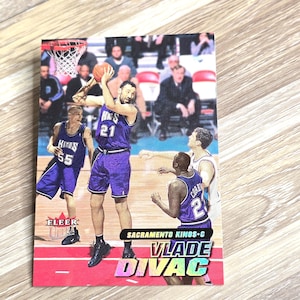May include: A Sacramento Kings basketball card featuring Vlade Divac in a purple uniform, mid-shot. The card includes the text "FLEER" and "SACRAMENTO KINGS-C VLADE DIVAC". The background shows other players and spectators.