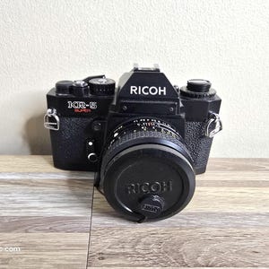 Peut inclure: Appareil photo argentique Ricoh KR-5 Super noir avec un bouchon d'objectif. Le nom de la marque "RICOH" est en lettres blanches sur le dessus et le bouchon de l'objectif. L'appareil photo est sur une surface en bois.