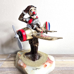 May include: A hand-painted figurine of a pilot with a red nose and mustache, wearing a brown flight suit and goggles, saluting while standing on a gold-toned airplane. The sculpture is mounted on a white stone base with the signature Romeros.