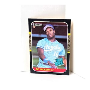 May include: A baseball card featuring Bo Jackson of the Kansas City Royals. The card has a black border with gold accents and the Donruss logo. Jackson is wearing a light blue Royals uniform with the number 16. The card reads "Rated Rookie".