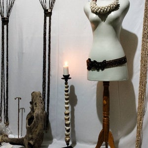 Peut inclure: Un mannequin blanc avec une ceinture en perles brunes et un collier de coquillages blancs. Le mannequin est sur un support en bois à trois pieds. Une bougie est allumée sur un chandelier à perles blanches et hautes. Le texte "Native Borneo Papua" est visible en bas de l'image.