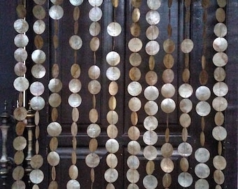 May include: A beaded curtain made of round, iridescent shell discs and oval beads hangs in front of a dark wooden door. The curtain is composed of multiple strands, creating a decorative and natural aesthetic. The text "Native Borneo Papua" is in the bottom right corner.