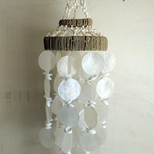 以下が含まれることがあります： 自然なデザインの吊り下げ式モビール。白い円形の貝殻のパーツが、ロープのような織り構造から吊り下げられています。上部は円錐形で、下部はカスケードデザインになっています。