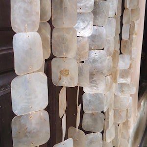May include: A decorative curtain made of square, iridescent capiz shell pieces strung together. The shells are a translucent white with subtle variations in color and texture. The curtain hangs vertically, creating a shimmering effect. The text "nativeborneopapua" is visible at the bottom.