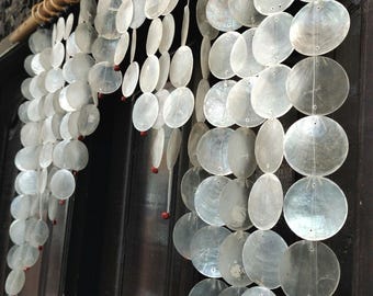 May include: A white wind chime made of seashells hangs from a wooden dowel. The shells are arranged in rows and are strung together with string. The wind chime is hanging in front of a dark wooden door.