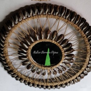 May include: A circular wall hanging made of woven natural fibres and decorated with small, brown and white seashells. The centre of the hanging features a round mirror with a black background and the text "Native Borneo Papua" in white.
