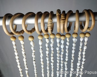 May include: A set of ten circular wooden curtain rings hanging on a silver rod, each featuring a dangling strand of white seashells attached by a small wooden bead. The text Native Borneo Papua is printed in white in the bottom right corner.