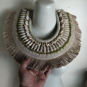 Puede incluir: Un collar grande hecho a mano con materiales naturales. El collar está hecho de una variedad de conchas, tejidas juntas con una fibra natural. El collar está adornado con una franja de la misma fibra natural. El collar es un estilo tradicional de Borneo, Papúa.