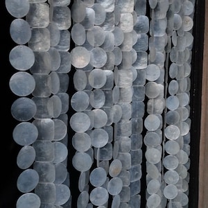 May include: A curtain made of multiple strands of white, round and square, iridescent, mother-of-pearl shells. The curtain is hanging in front of a dark background. The text "Native Borneo Papua" is visible at the bottom of the image.