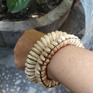 Puede incluir: Un brazalete de hueso blanco con un diseño tejido. El brazalete se lleva en la muñeca de una persona. El texto "Native Borneo Papua" es visible en la parte inferior de la imagen.