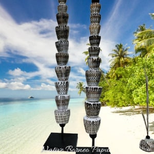 Puede incluir: Dos esculturas altas hechas de conchas apiladas con patrones blancos y negros sobre soportes negros. Las esculturas están en una playa de arena blanca con palmeras y un cielo azul de fondo. "Native Borneo Papua" está escrito en la base de uno de los soportes.