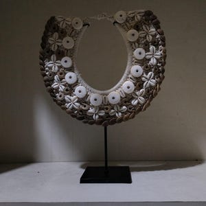 May include: A shell necklace with a U-shaped design, displayed on a black metal stand. The necklace is decorated with various seashells and white circular accents, showcasing the intricate craftsmanship and design.