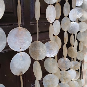 Puede incluir: Cortina decorativa hecha de conchas capiz redondas y ovaladas iridiscentes. Las conchas están ensartadas con un hilo fino y casi invisible, creando un efecto brillante. La cortina cuelga frente a una puerta de madera marrón oscuro.