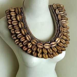 May include: A handcrafted necklace made of numerous brown seashells, arranged in a layered design. The shells are attached to a woven, rope-like base, creating a statement piece. The necklace is displayed on a white mannequin.