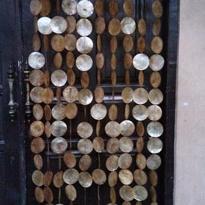 Könnte beinhalten: Ein dekorativer Türvorhang aus runden, irisierenden, natürlichen Muschelstücken, die aneinandergereiht sind. Der Vorhang hängt vor einer dunklen Holztür. Die Muschelstücke sind in verschiedenen Brauntönen und Weiß gehalten.