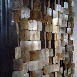 Peut inclure: Un rideau décoratif composé de morceaux de coquillages carrés et rectangulaires dans des tons de marron, beige et blanc. Les coquillages sont enfilés ensemble, créant un aspect texturé et naturel. Le texte "Native Borneo Papua" est visible.