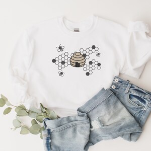 May include: White sweatshirt with a black and brown beehive design. The beehive is surrounded by black honeycomb shapes and small black bees.
