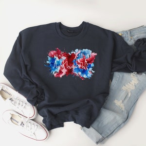 Puede incluir: Sudadera azul marino con un diseño de ciudad blanca sobre un fondo de acuarela rojo, blanco y azul.