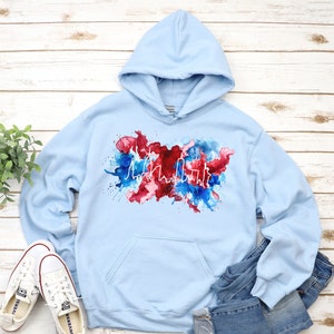 Puede incluir: Sudadera con capucha azul claro con un contorno blanco de un horizonte de ciudad sobre un fondo de acuarela rojo, blanco y azul.