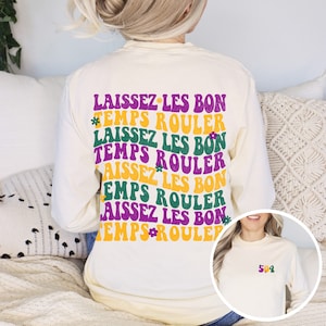 Puede incluir: Una camiseta de manga larga de color crema con la frase "Laissez Les Bon Temps Rouler" repetida en texto morado, verde y amarillo. La camiseta tiene un diseño de flor al lado del texto. Una segunda imagen muestra una camiseta crema con el número "502" en texto morado, verde y amarillo.