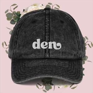 May include: A black distressed denim baseball cap with white embroidered text that reads "deno".