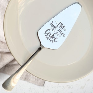 Pode incluir: Um servidor de bolo de prata com uma borda serrilhada e uma alça. O servidor tem o texto "I'm Just Here for the Cake" gravado.