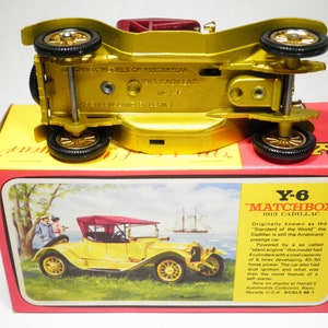 May include: A gold Matchbox Y-6 1913 Cadillac toy car model. The car is on a red and yellow box with a picture of the car and text that reads "Originally known as the 'Standard of the World' the Cadillac is still the Americans prestige car."
