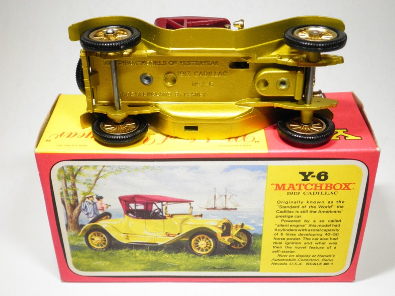 May include: A gold Matchbox Y-6 1913 Cadillac toy car model. The car is on a red and yellow box with a picture of the car and text that reads "Originally known as the 'Standard of the World' the Cadillac is still the Americans prestige car."