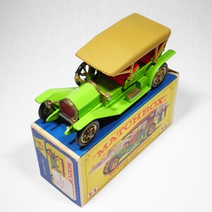 May include: A green toy car with a tan roof and gold wheels. The car is sitting on a blue and yellow box with the words "Matchbox" and "Models of Yesteryear" printed on it.