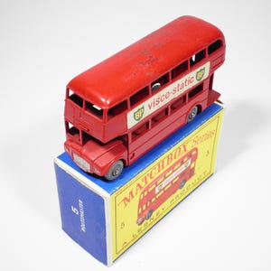 May include: A red double-decker bus toy with the text "BP visco-static" on the side, sitting on a blue and yellow box. The box is labeled "MATCHBOX Series" and features a bus illustration.