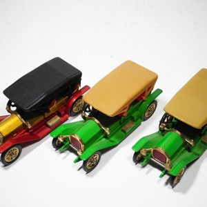 May include: Three vintage toy cars, one red, one green, and one green with a tan top. The cars have gold accents and black wheels.