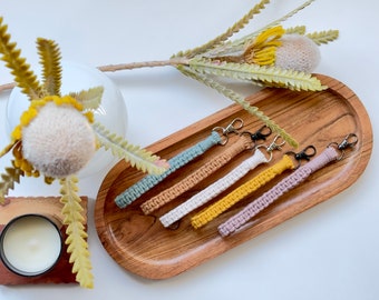 May include: Six crocheted keychains in various colors, including teal, beige, white, yellow, pink, and brown, are displayed on a wooden tray. Each keychain has a black metal ring.