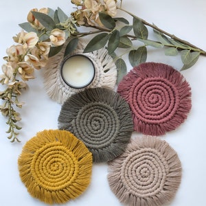 May include: Five handmade macrame coasters in different colors, including beige, gray, pink, and yellow. The coasters have a circular shape with a fringe border and a spiral design in the center.