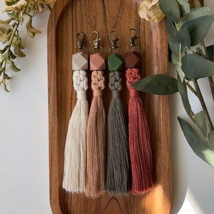 May include: Four keychains with wooden beads and tassels. The beads are in shades of beige, pink, green, and brown. The tassels are in matching colors and are made of a soft, fluffy material.