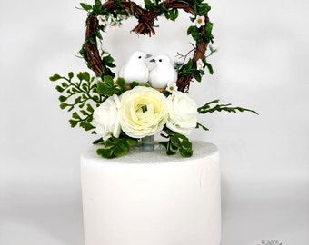 May include: White cake topper with two white birds perched inside a heart-shaped twig wreath with white flowers. The wreath is surrounded by white flowers and green leaves.