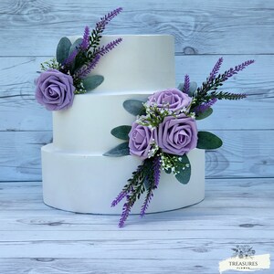 May include: A three-tiered white wedding cake decorated with purple roses and lavender sprigs.