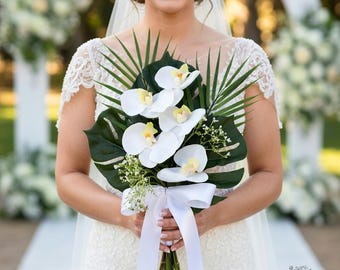 May include: A bride holding a bouquet of white orchids with yellow centers, green leaves, and a white ribbon. The bouquet includes large, dark green leaves and small white flowers. The bride is wearing a white lace dress and veil.