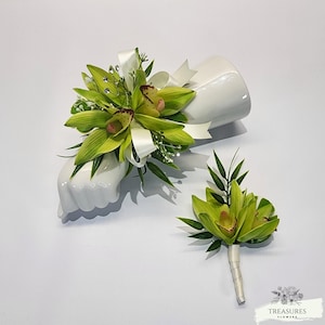 May include: A green orchid corsage and boutonniere set with white satin ribbon and rhinestones. The corsage is displayed on a white ceramic hand.