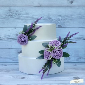 May include: A three-tiered white wedding cake decorated with lavender and purple roses and greenery. The cake is on a wooden background.