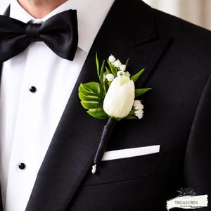 May include: A white tulip boutonniere with green leaves and small white flowers, attached to a black stem, pinned to a black tuxedo jacket. The jacket has a white pocket square and a black bow tie.