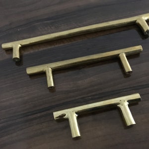 May include: Three gold-toned metal cabinet pulls of different sizes. The pulls are T-shaped with a straight bar and a perpendicular handle.