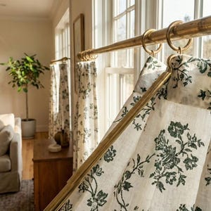 May include: Cream-colored floral curtains hanging from a gold-toned rod. The curtains feature a dark green floral pattern. The room has a neutral color palette with a sofa, wooden furniture, and a potted plant.